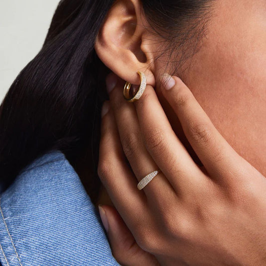Close-up of a person wearing gold hoop earrings and a ring, with a neutral background.