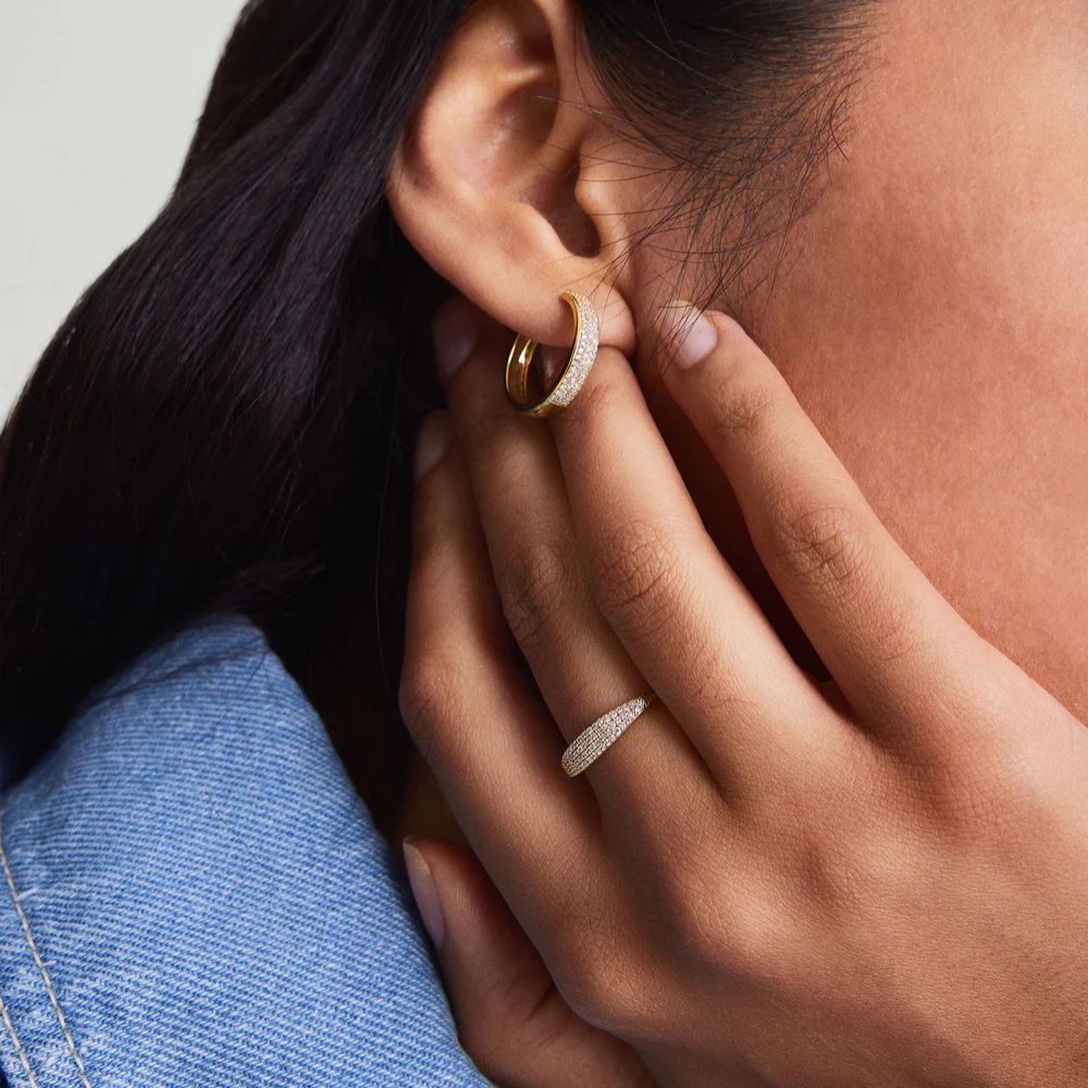 Close-up of a person wearing gold hoop earrings and a ring, with a neutral background.